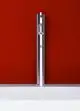Chromfarbenes Standventil von Ideal Standard, vertikal montiert, auf einer weißen Fläche, vor roter Wand. Funktionales Design ohne sichtbare Maße oder Details.